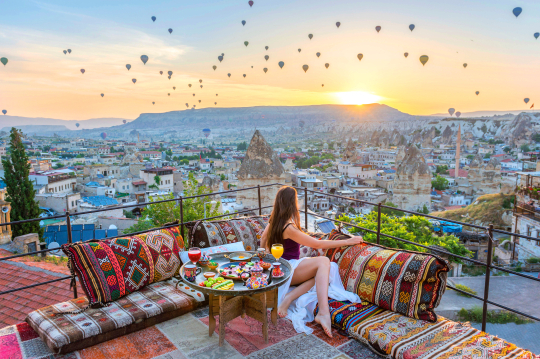 Frau sitzt auf einer Hotelterrasse mit Blick über die Landschaft von Kappadokien und bestaunt die aufsteigenden Heißluftballons